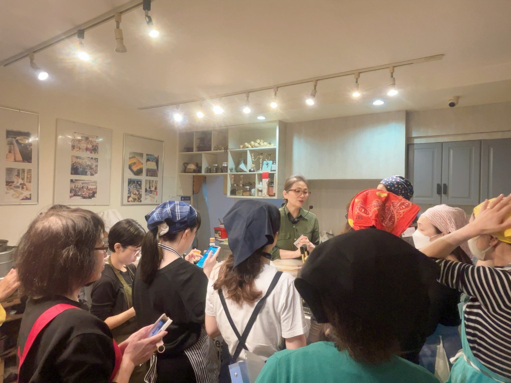 料理教室で講師が参加者に説明している様子。参加者はエプロンやバンダナを着用している。