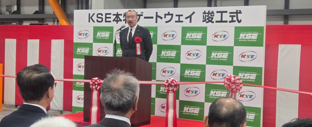 KSE本社の開設式でスピーチをする男性。後ろには観衆が集まり、紅白のリボンとKSEのロゴが描かれたバナーが見える。
