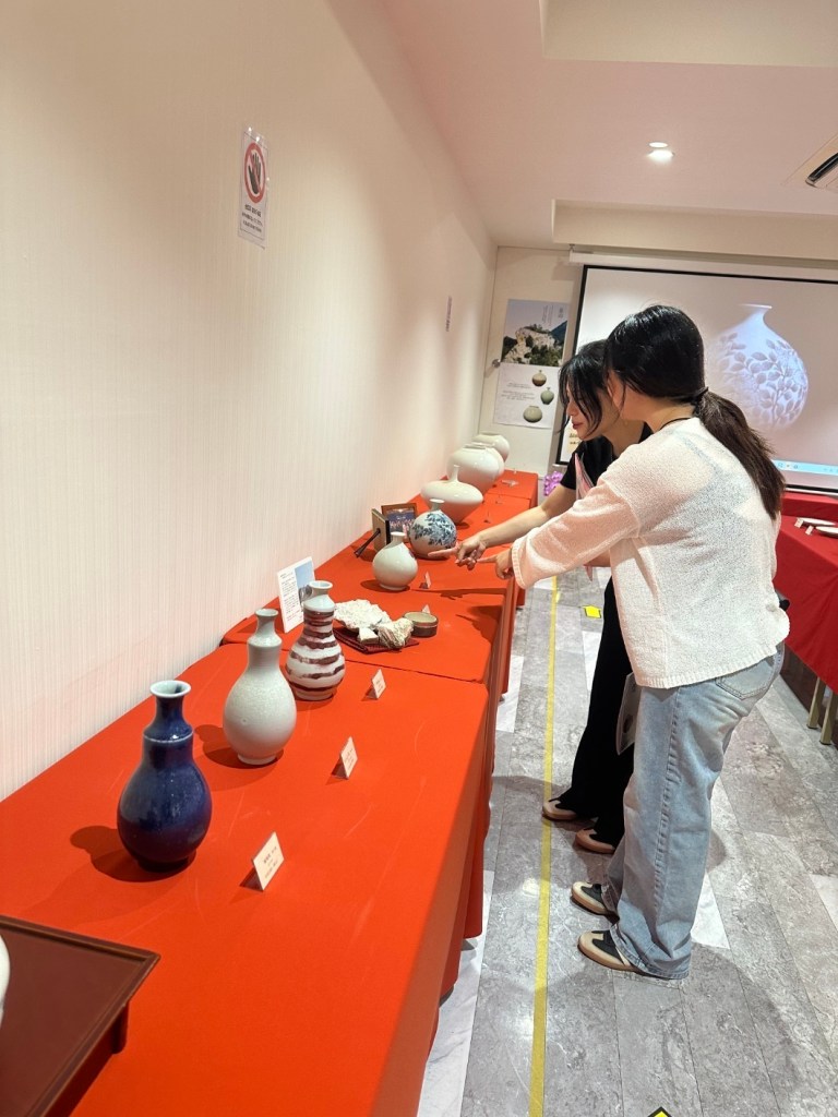展示された陶器を観賞する二人の女性。赤いテーブルクロスの上に様々な形やデザインの陶器が並んでいる。