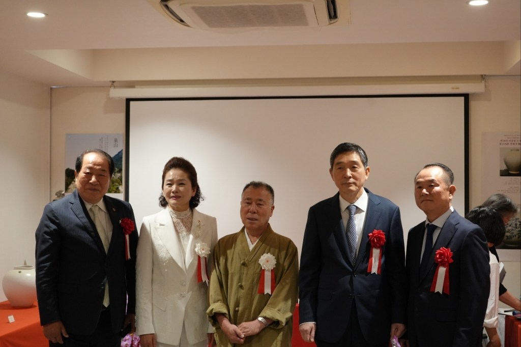公式イベントで並んで立つ五人の男性と女性。背景にはスクリーンがあり、赤いリボンを付けた服装をしています。